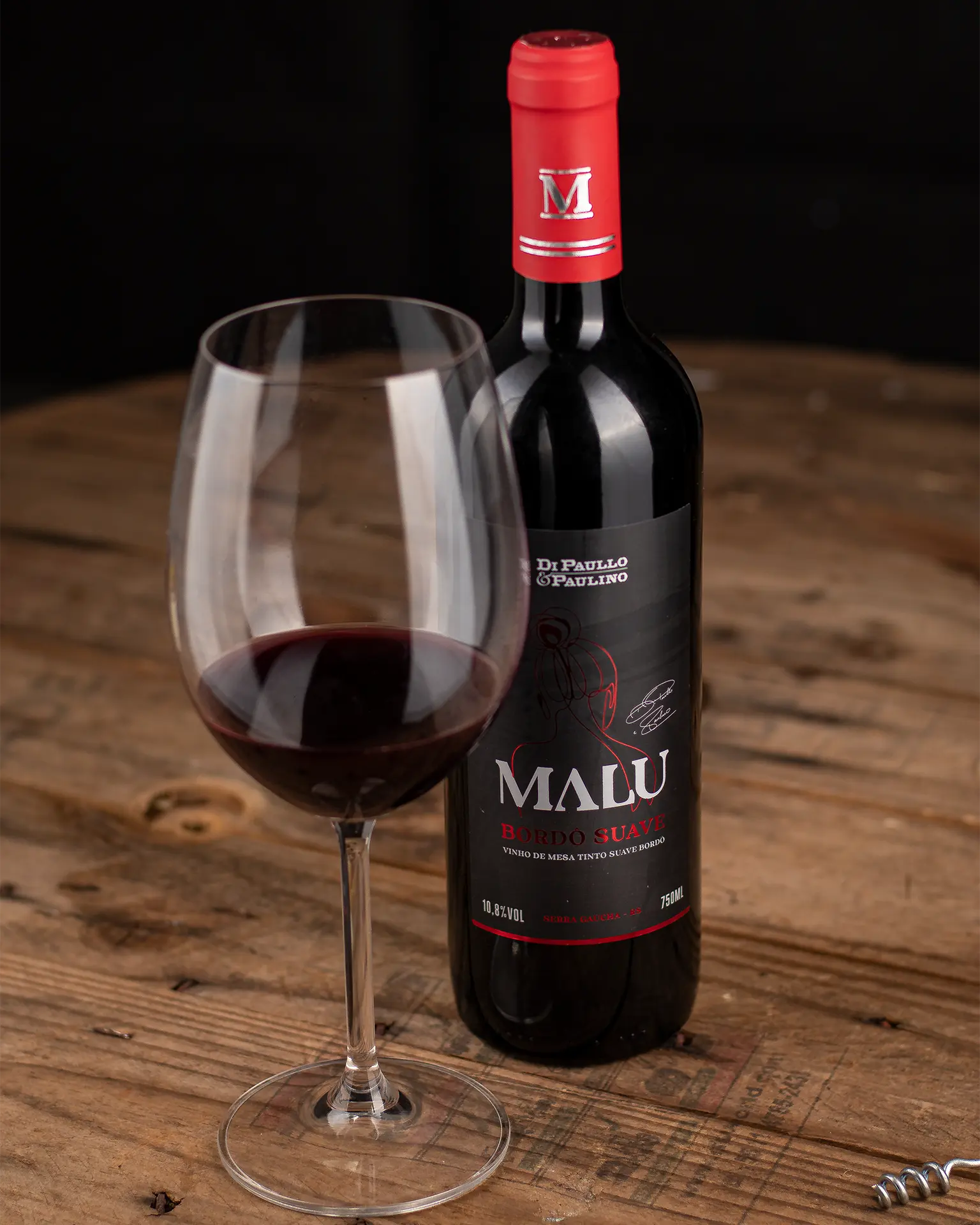 Garrafa de vinho de mesa Malu Bordô Suave ao lado de uma taça